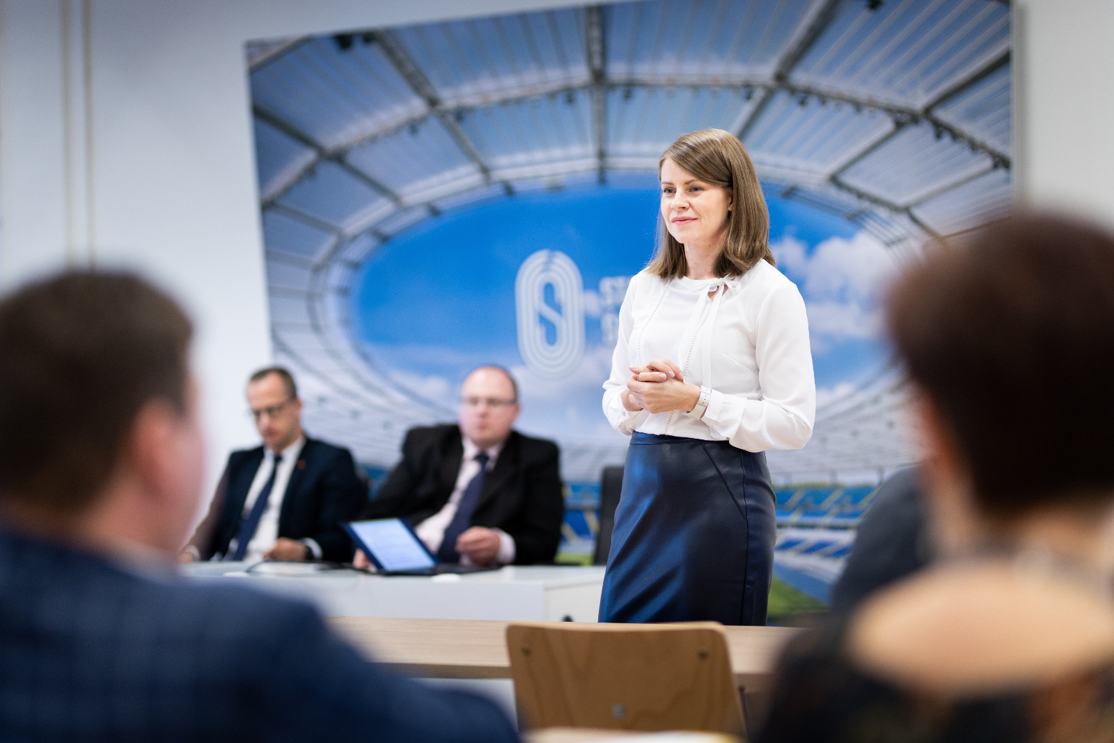 Konferencja Stadion Śląski - Kontraktacja i rozliczanie. Zrób to dobrze: Na pierwszym planie kobieta w białej bluzce i granatowej spódnicy prowadzi warsztat pod tytułem Kontraktacja i rozliczanie. Zrób to dobrze. W tle za kobietą dwóch mężczyzn w garniturach siedzących za stołem konferencyjnym.