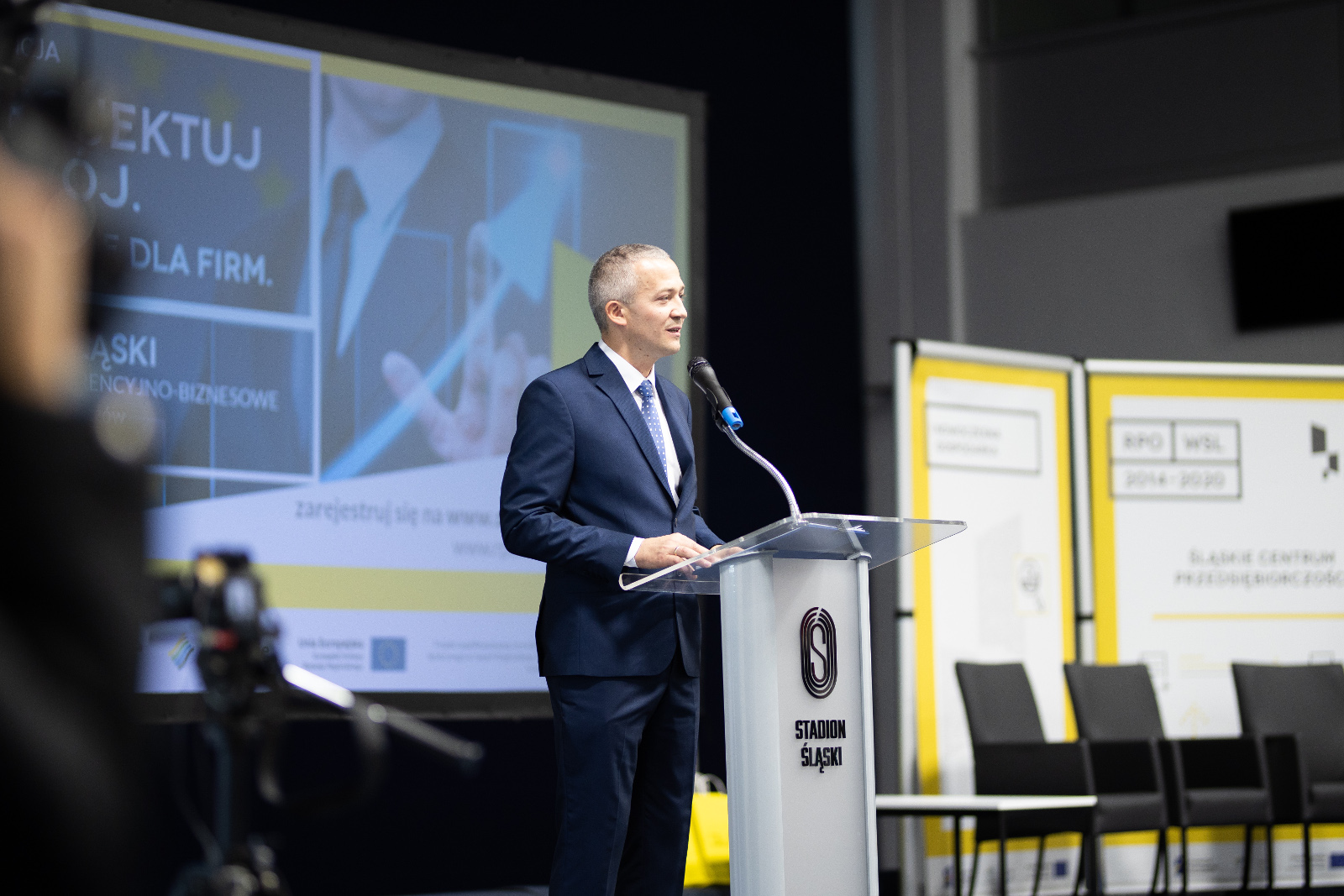 Konferencja Stadion Śląski - Krzysztof Spyra, Dyrektor Śląskiego Centrum Przedsiębiorczości: Mężczyzna w granatowym garniturze wita gości przy pulpicie konferencyjnym. W tle ekran wyświetlający grafikę konferencji Zaprojektuj rozwój. Wsparcie UE dla firm oraz fotele ustawione do zaplanowanej dyskusji panelowej.