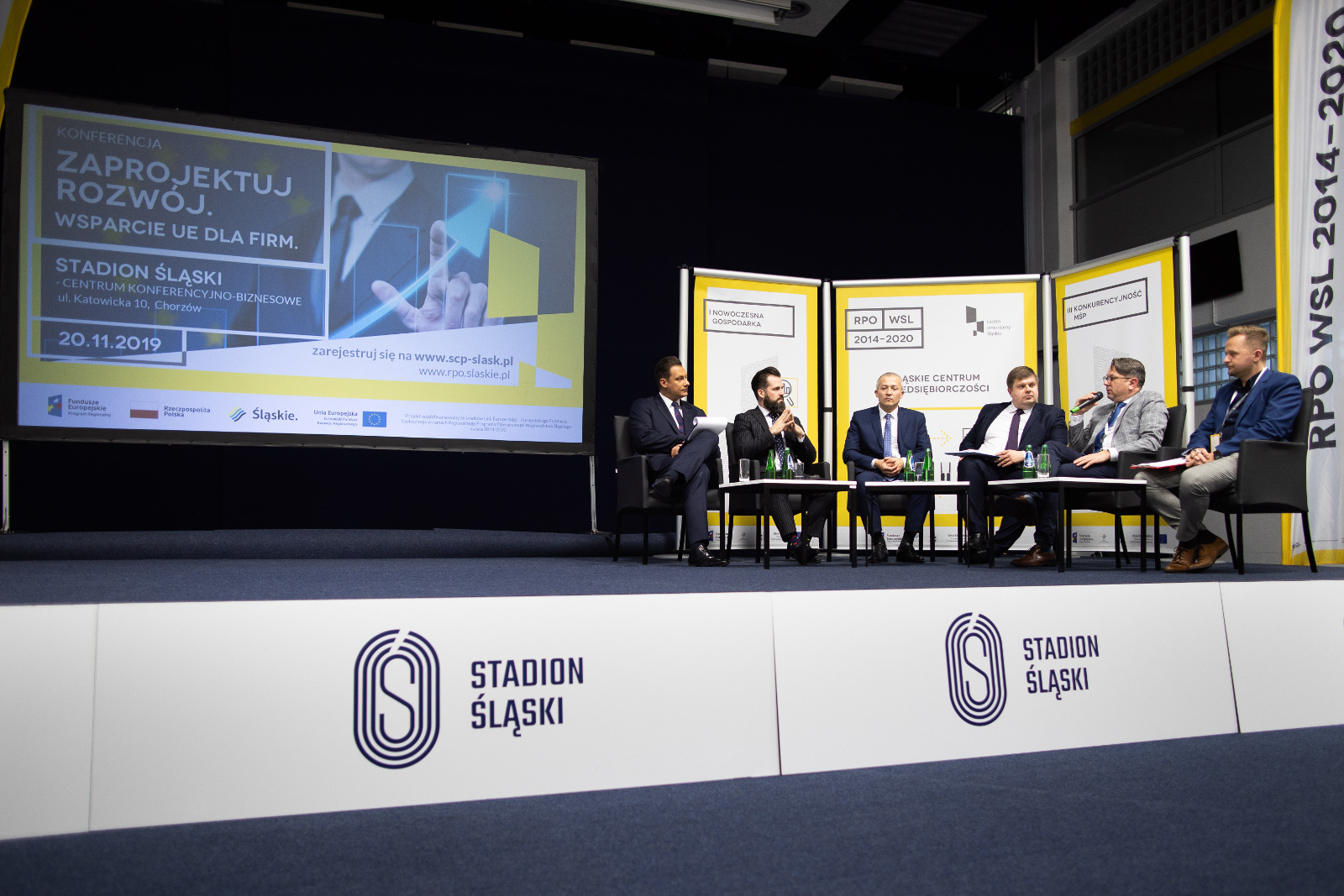 Konferencja Stadion Śląski - dyskusja panelowa: Na zdjęciu 6 mężczyzn w garniturach uczestniczy w dyskusji panelowej. Siedzą w czarnych fotelach rozmieszczonych w półkolu na tle biało żółtej ścianki wystawienniczej Śląskiego Centrum Przedsiębiorczości. Tło do zdjęcia stanowi ekran wyświetlający grafikę konferencji Zaprojektuj rozwój. Wsparcie UE dla firm. Dyskusja panelowa prowadzona jest na podwyższonej scenie opatrzonej banerami Stadion Śląski wskazującymi na miejsce realizacji konferencji