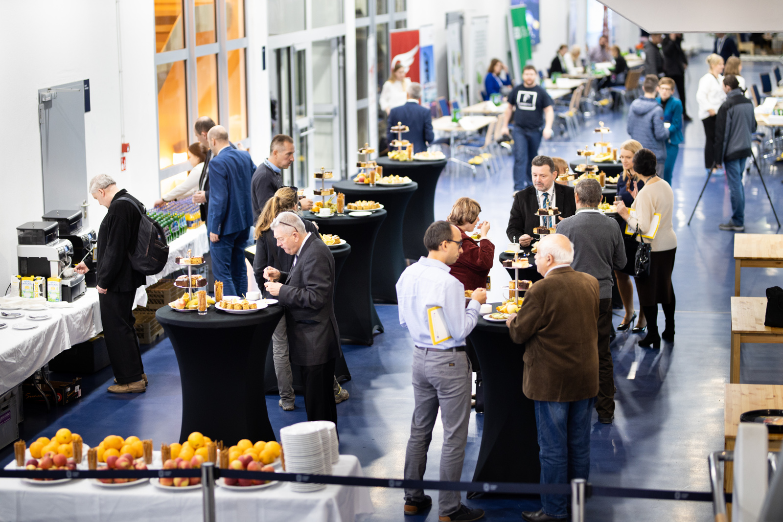 Konferencja Stadion Śląski - stanowiska informacyjne: Widok z góry na catering oraz 15 stanowisk informacyjnych dostępnych podczas konferencji. Na pierwszym planie talerze z owocami. W dalszej części zdjęcia ludzie stojący przy wysokich stołach bankietowych. Po prawej stronie zdjęcia niskie stoliki dedykowane dla osób poruszających się na wózkach inwalidzkich. Po lewej stronie zdjęcia wzdłuż ścian ekspresy do kawy.