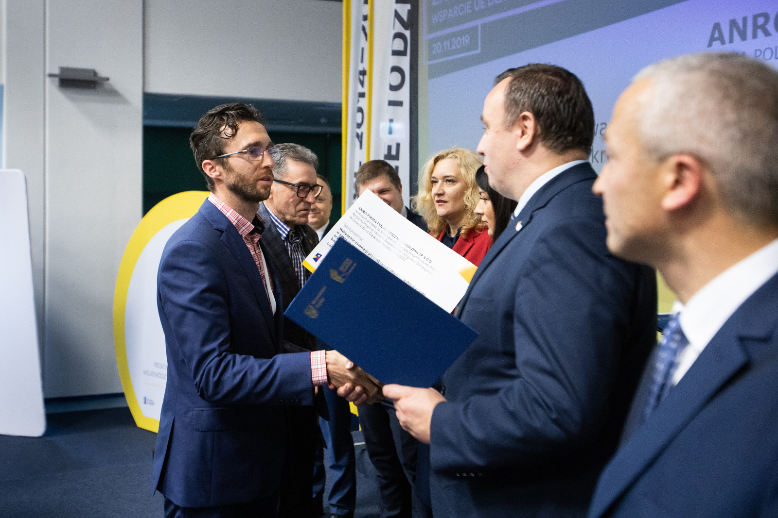 Konferencja Stadion Śląski - uroczyste wręczenie umów o dofinansowanie - ANRO: Na zdjęciu widoczni: Dyrektor Śląskiego Centrum Przedsiębiorczości oraz Przedstawiciele Zarządu Województwa  trzech mężczyzn i dwie kobiety wręczają umowę o dofinansowanie dwóm przedstawicielom firmy ANRO. Marszałek Województwa Śląskiego Jakub Chełstowski, ściskając dłoń Przedsiębiorcy przekazuje umowę oprawioną w elegancką niebieską teczkę. Mężczyźni ubrani w granatowe garnitury. 