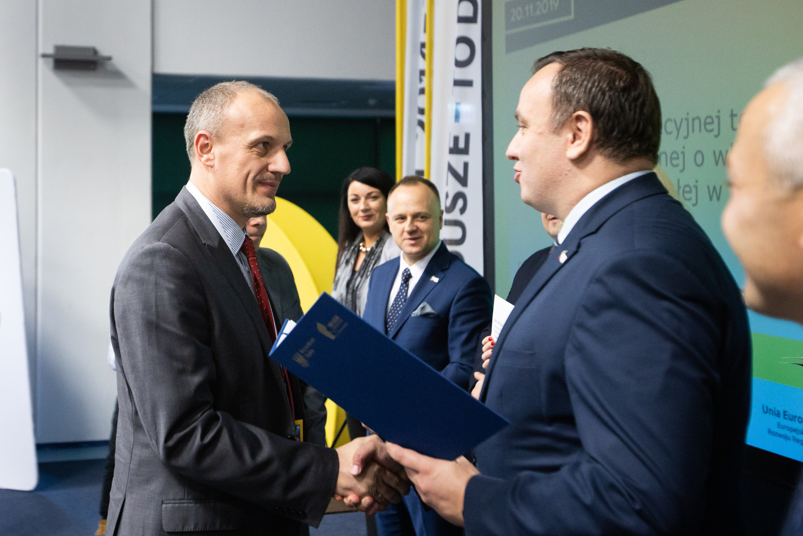 Konferencja Stadion Śląski - uroczyste wręczenie umów o dofinansowanie - ERG: Na pierwszym planie Marszałek Województwa Śląskiego Jakub Chełstowski, w granatowym garniturze, ściskając dłoń Przedstawiciela firmy ERG (w szarym garniturze), przekazuje umowę oprawioną w elegancką niebieską teczkę. Na zdjęciu widoczni również - Dyrektor Śląskiego Centrum Przedsiębiorczości Krzysztof Spyra oraz Przedstawiciele Zarządu Województwa: Dariusz Starzycki (w niebieskim garniturze) oraz Izabela Domogała ubrana w szarą marynarkę i szary, wzorzysty szal.