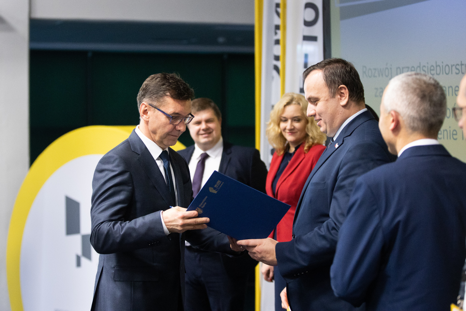 Konferencja Stadion Śląski - uroczyste wręczenie umów o dofinansowanie - MCJ: Na pierwszym planie Marszałek Województwa Śląskiego Jakub Chełstowski, ściskając dłoń Przedstawiciela firmy MCJ, przekazuje umowę oprawioną w elegancką niebieską teczkę. Na zdjęciu widoczni również - Dyrektor Śląskiego Centrum Przedsiębiorczości Krzysztof Spyra oraz Przedstawiciele Zarządu Województwa: Wojciech Kałuża (w niebieskim garniturze) oraz Beata Białowąs ubrana w czerwoną marynarkę i granatową sukienkę.