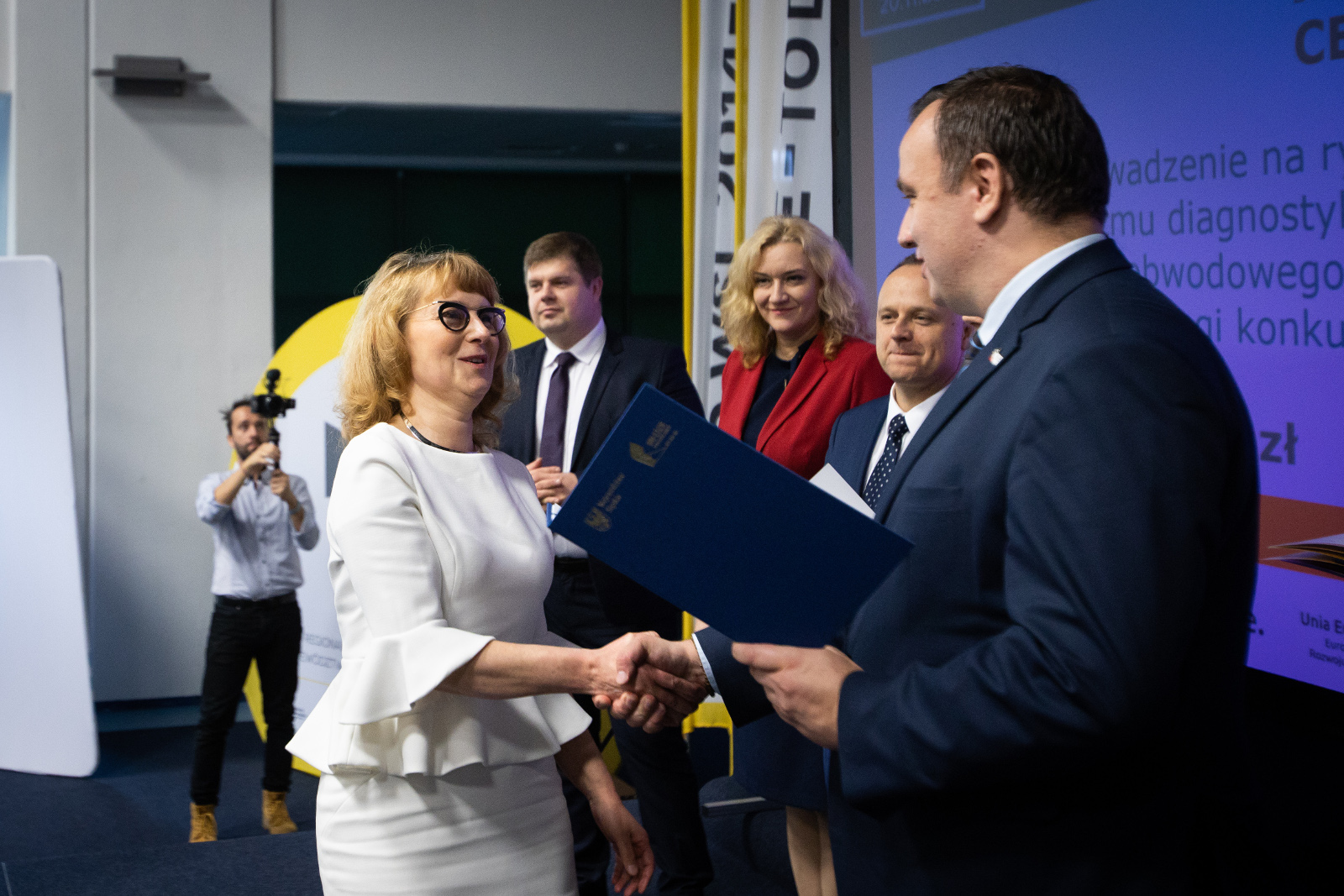 Konferencja Stadion Śląski - uroczyste wręczenie umów o dofinansowanie - Sanvita: Na pierwszym planie Marszałek Województwa Śląskiego Jakub Chełstowski, ściskając dłoń Przedstawicielki firmy SANVITA (w białej sukience i czarnych oprawkach okularowych), przekazuje umowę oprawioną w elegancką niebieską teczkę. Na zdjęciu widoczni również - Centrum Przedstawiciele Zarządu Województwa: Wojciech Kałuża (w niebieskim garniturze), Beata Białowąs ubrana w czerwoną marynarkę i granatową sukienkę oraz Dariusz Starzycki w granatowym garniturze. W drugim planie, z boku sceny widoczny fotograf. 