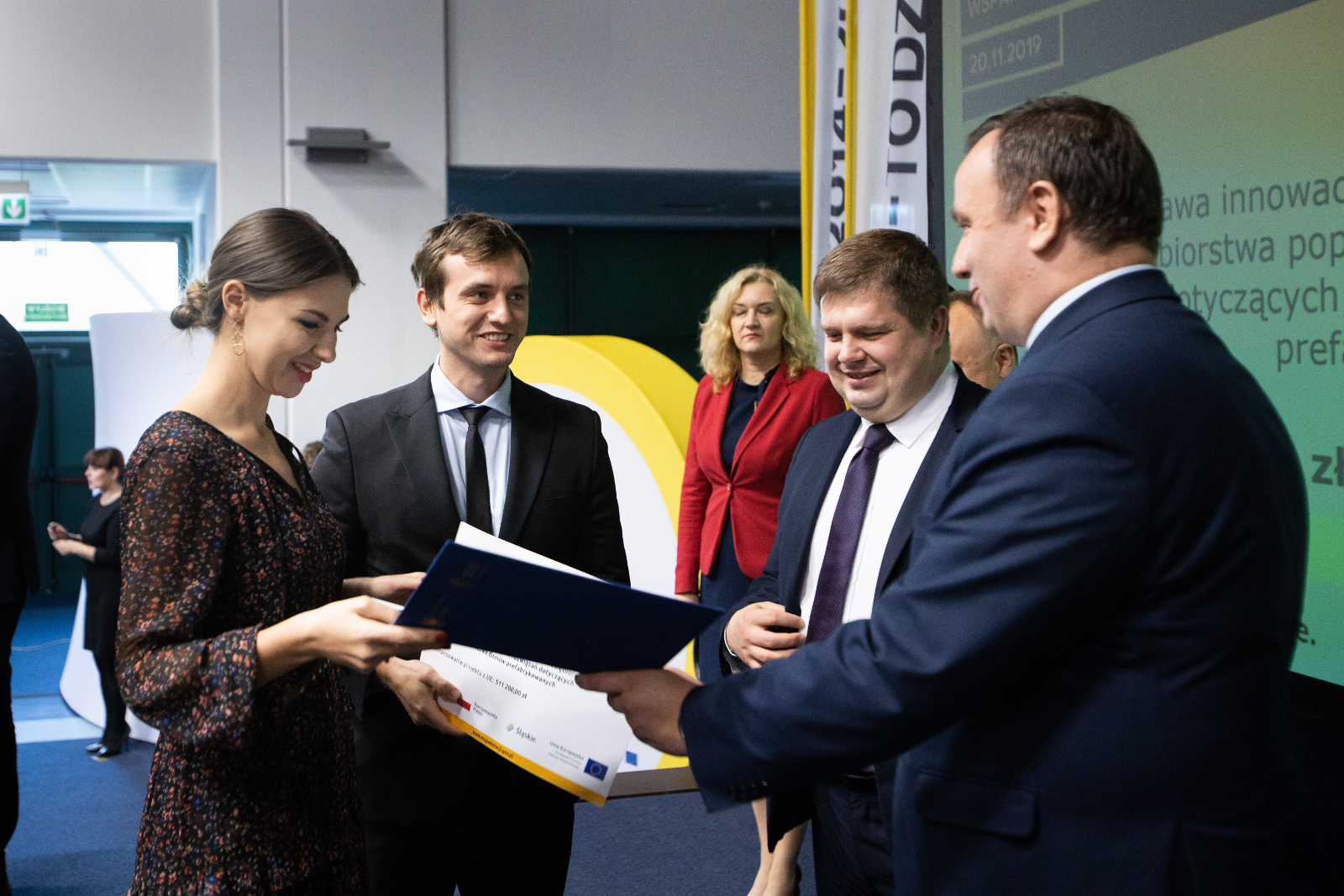 Konferencja Stadion Śląski - uroczyste wręczenie umów o dofinansowanie - Tadeks Fertig Haus: Na pierwszym planie Marszałek Województwa Śląskiego Jakub Chełstowski przekazuje umowę oprawioną w elegancką niebieską teczkę Przedstawicielom firmy Tadeks Fertig Haus. Kobieta w brązowej sukience w drobne kwiaty odbiera umowę, mężczyzna w czarnym garniturze trzyma pamiątkowy plakat informacyjny w zakresie realizowanego projektu. Na zdjęciu widoczni również - Przedstawiciele Zarządu Województwa: Wojciech Kałuża oraz Beata Białowąs. Na drugim planie zdjęcia widoczna tłumacz migowa relacjonująca wydarzenia na scenie.