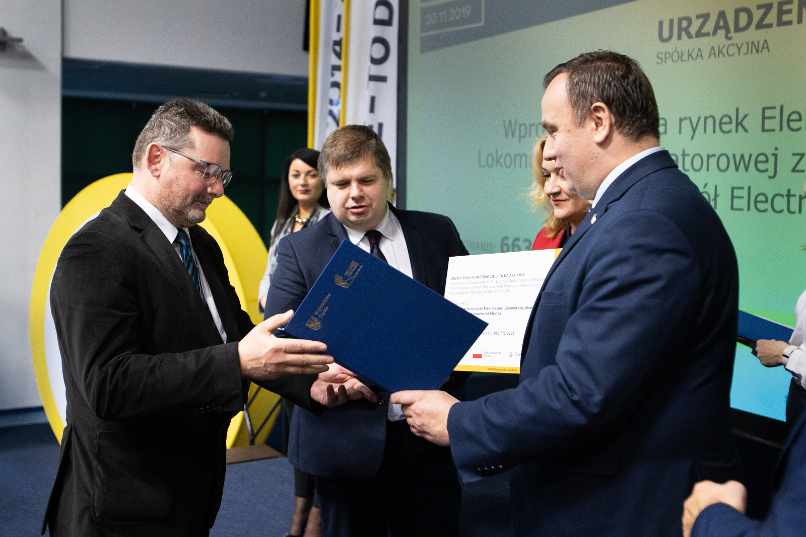 Konferencja Stadion Śląski - uroczyste wręczenie umów o dofinansowanie - URZĄDZENIA I KONSTRUKCJE: Na pierwszym planie Marszałek Województwa Śląskiego Jakub Chełstowski przekazuje umowę oprawioną w elegancką niebieską teczkę Przedstawicielowi firmy Urządzenia i Konstrukcje ubranemu w ciemnobrązową marynarkę. Na zdjęciu widoczna również Członek Zarządu Województwa Beata Białowąs, która wręcza  pamiątkowy plakat informacyjny w zakresie realizowanego projektu, a także Wojciech Kałuża Wicemarszałek Województwa. Na drugim planie zdjęcia widoczna Członek Zarządu Izabela Domogała