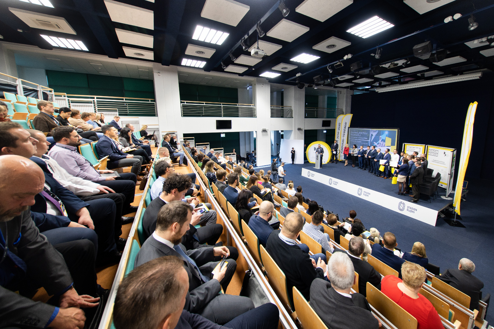 Konferencja Stadion Śląski - widok z sali na uroczyste wręczenie umów o dofinansowanie: Na zdjęciu uczestnicy konferencji zajmujący miejsca na Sali w ustawieniu amfiteatralnym. U dołu zdjęcia scena, na której stoją wszyscy przedsiębiorcy, po uroczystości wręczenia umowy o dofinansowanie. Jeden z przedsiębiorców przy pulpicie zabiera glos.